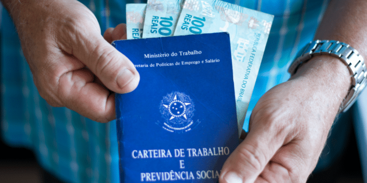 Mãos seguram Carteira de Trabalho azul e notas de 100 reais em roupa xadrez azul.