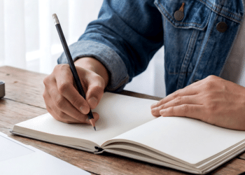 Pessoa escreve em caderno aberto com lápis preto em ambiente de trabalho.