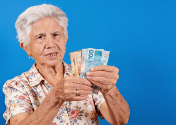 Idosa exibe notas de real em fundo azul com expressão confiante.