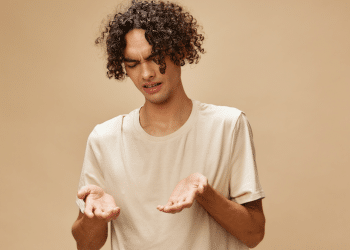 Jovem confuso observa as próprias mãos em fundo bege.