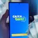 Mão segurando celular com tela do app Caixa Tem aberta e notas de cem reais ao fundo, simbolizando a movimentação do dinheiro do Pé-de-Meia pela conta Caixa Tem.