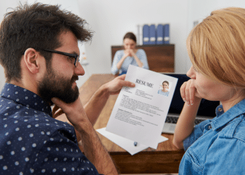 Homem mostra currículo impresso para mulher enquanto recrutadora observa ao fundo em escritório.