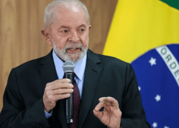 Lula segurando microfone em frente à bandeira brasileira, fazendo gesto com a mão.