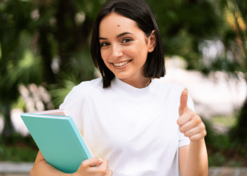 Jovem mulher segura cadernos coloridos e mostra joinha, sorrindo em ambiente externo verde, simbolizando o processo seletivo com vagas para Assistente Social.
