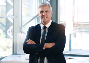 Homem de terno e com os braços cruzados no seu ambiente de trabalho.