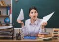 Mulher jovem de cabelos escuros rasga papel ao meio, sentada à mesa com livros, ábaco e quadro-negro ao fundo, simbolizando a dúvida sobre o aumentativo de papel.