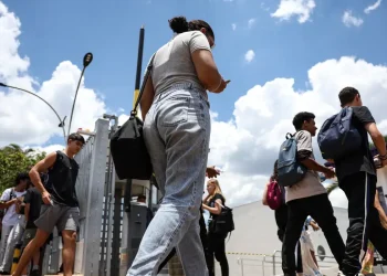 Candidatos com mochilas atravessam o portão de entrada do local de prova do Enem em dia ensolarado