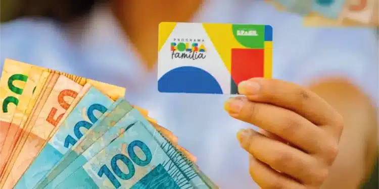 Beneficiária segura cartão do Bolsa Família e notas de real durante o calendário de pagamentos de julho.