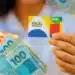 Beneficiária segura cartão do Bolsa Família e notas de real durante o calendário de pagamentos de julho.