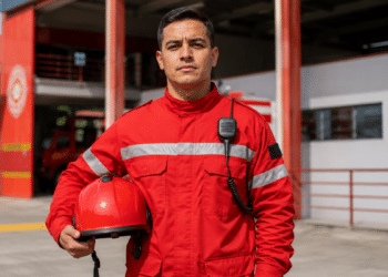 Imagem ilustrativa de bombeiro civil de uniforme vermelho e rádio de comunicação em frente a um quartel, representando a profissão amparada por lei federal