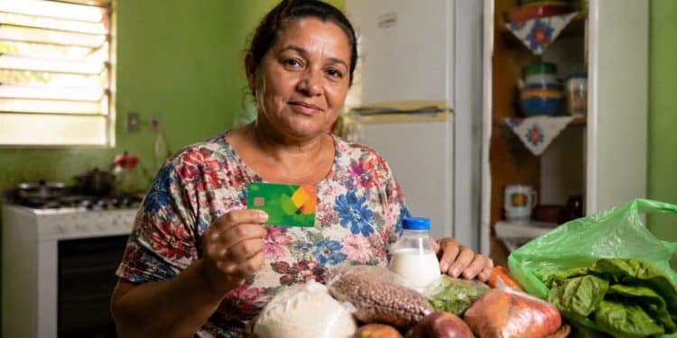 Mulher segura um cartão alimentação ao lado de uma cesta com arroz, feijão, frutas, legumes e leite, ilustrando os programas estaduais que pagam de R$ 100 a R$ 300 por mês