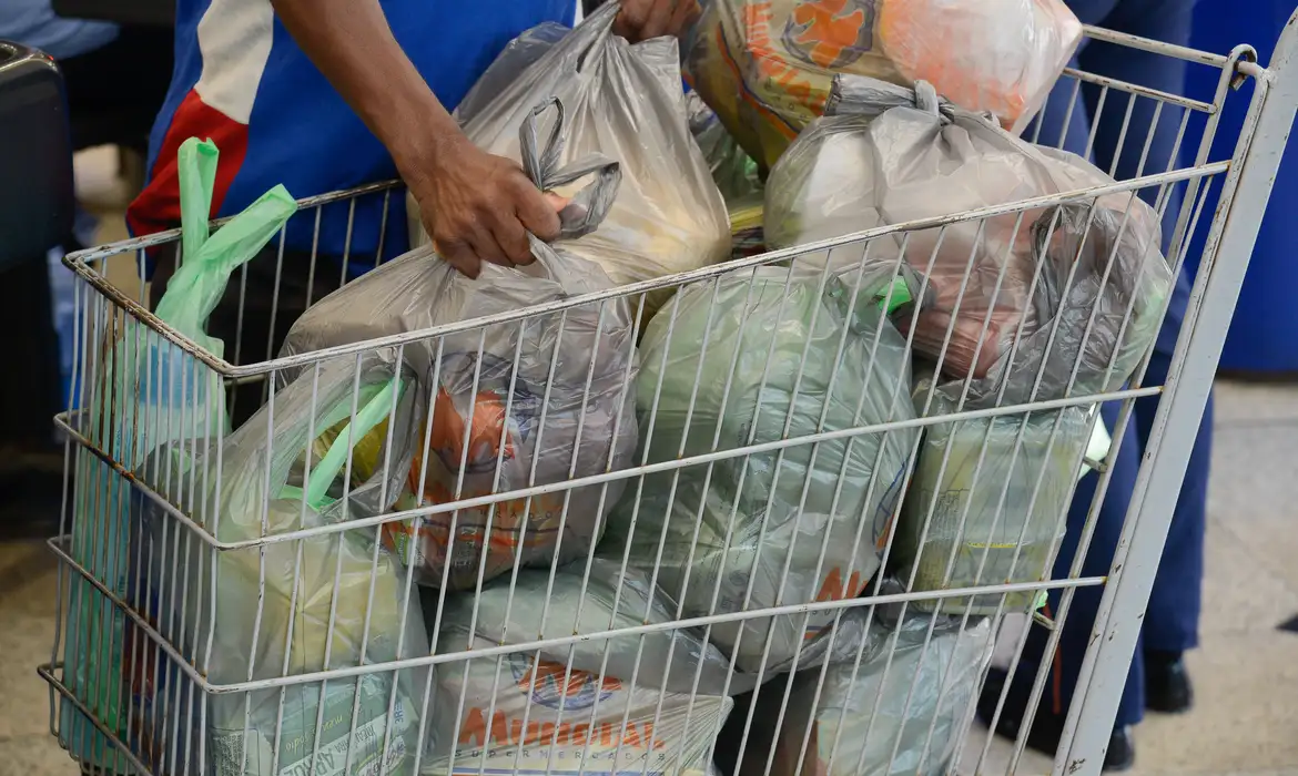 Homem coloca sacolas de compras de supermercado em um carrinho, ilustrando o uso do cartão alimentação para a compra de mantimentos