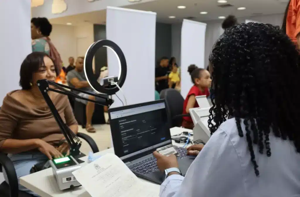 Imagem de atendente coletando a biometria de uma cidadã em um posto de identificação, com leitor de digital e câmera sobre a mesa