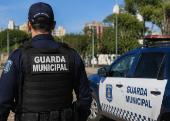 Guarda Municipal de costas em patrulhamento ao lado de viatura oficial em área urbana.
