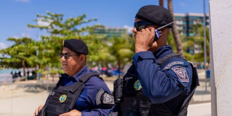 Imagem ilustrativa de dois agentes de guarda municipal em serviço na via pública, de farda azul-marinho, colete tático e boina, um deles falando ao rádio comunicador