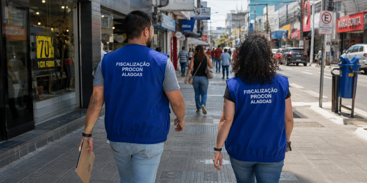 Dois fiscais do Procon de Alagoas caminham por uma rua comercial durante atividade de fiscalização.