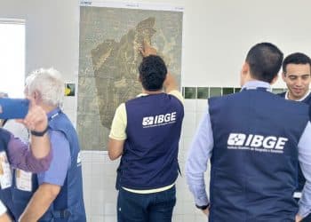 Servidores com colete do IBGE analisam mapa de município fixado na parede durante trabalho de campo