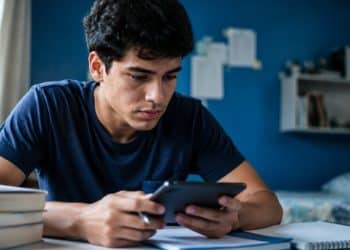 Estudante consulta o celular à mesa, com livros e caderno, para verificar informações do Encceja 2026 na Página do Participante.