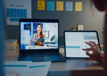 Jovem ministra aula online em notebook enquanto pessoa analisa gráficos em tablet no escritório, simbolizando os cursos livres para o currículo.