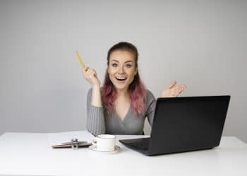 Mulher jovem sorridente com cabelo rosa segura lápis ao lado de notebook em mesa branca, simbolizando as opções de curso para ganhar dinheiro online.