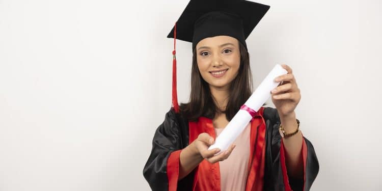 Jovem estudante sorrindo com beca preta e detalhes vermelhos segura diploma enrolado em destaque, simbolizando o prazo final para pedir atendimento especializado no Enade 2026.