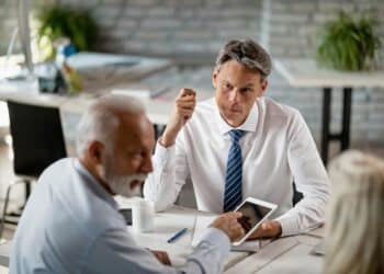 Imagem ilustrativa de homem de camisa branca e gravata conversando com um casal do outro lado da mesa em uma reunião de trabalho, representação da negociação de uma proposta de emprego