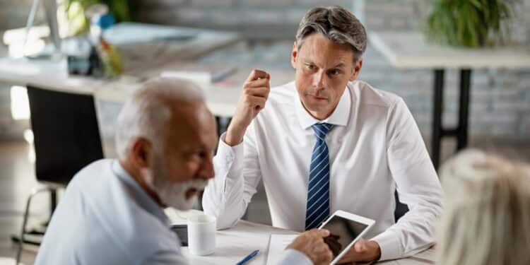 Imagem ilustrativa de homem de camisa branca e gravata conversando com um casal do outro lado da mesa em uma reunião de trabalho, representação da negociação de uma proposta de emprego
