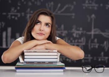 Jovem mulher apoia o queixo nos braços sobre pilha de cadernos diante de quadro negro com fórmulas matemáticas, simbolizando a dúvida entre esperto e experto.
