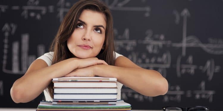 Jovem mulher apoia o queixo nos braços sobre pilha de cadernos diante de quadro negro com fórmulas matemáticas, simbolizando a dúvida entre esperto e experto.