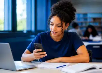 Jovem universitária consulta informações sobre a inscrição no Fies pelo celular, com notebook e documentos sobre a mesa.