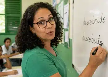 Professora escrevendo “descoberto” no quadro em sala de aula, representando a forma correta entre “descoberto” e “descobrido”.