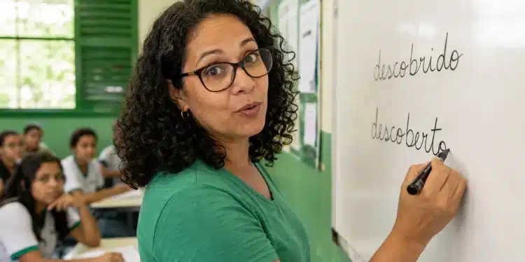 Professora escrevendo “descoberto” no quadro em sala de aula, representando a forma correta entre “descoberto” e “descobrido”.