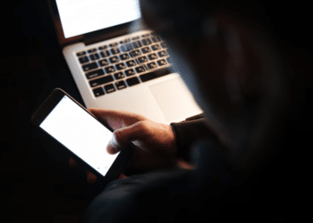 Pessoa segurando um celular iluminado diante de um notebook em ambiente escuro