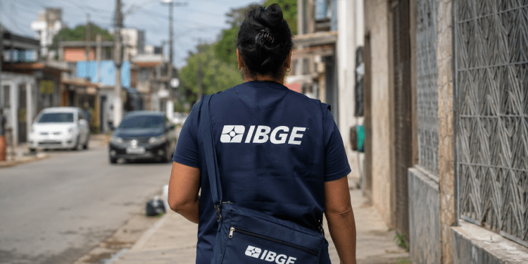 Pessoa uniformizada do IBGE caminha de costas em rua residencial durante trabalho de campo.