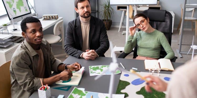 Equipe multidisciplinar analisa mapas e gráficos ao lado de maquetes de turbinas eólicas em reunião de projeto ambiental