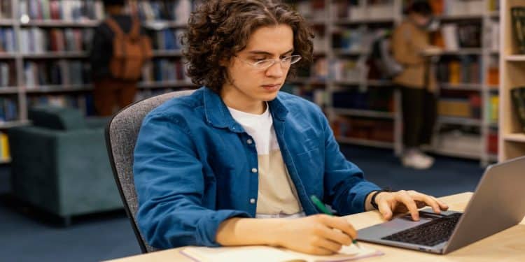 Estudante de óculos escreve em caderno diante do notebook, montando a rotina de revisão descrita na matéria.