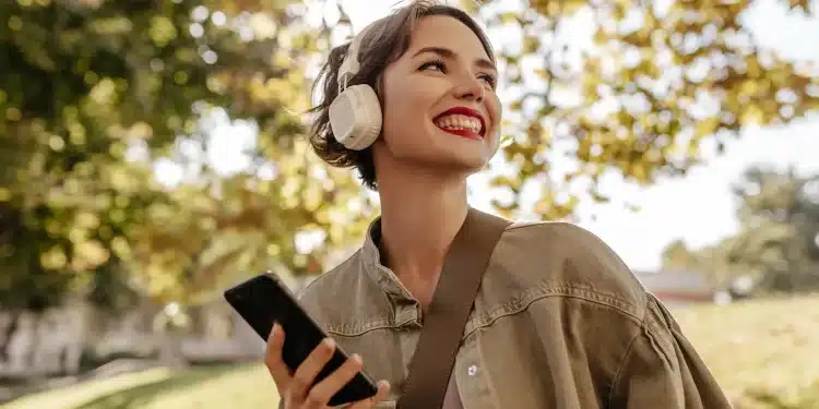 Mulher sorridente ouve música com fones de ouvido enquanto segura o celular em um parque.