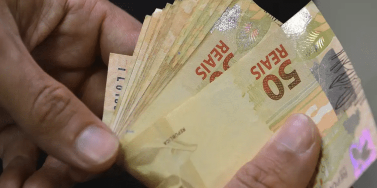 Mãos segurando várias notas de 50 reais em leque, fundo escuro, simbolizando o pagamento do INSS no mês de agosto.