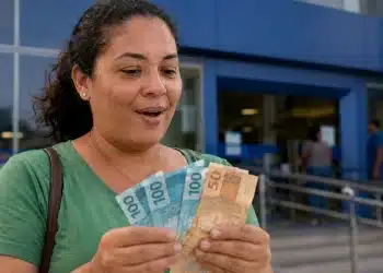 Mulher segurando cédulas de real em frente a uma entrada azul, representando pagamentos extras liberados em julho.