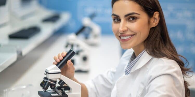 Imagem ilustrativa de profissional de jaleco branco ao lado de um microscópio em laboratório, em referência à profissão de biomédico