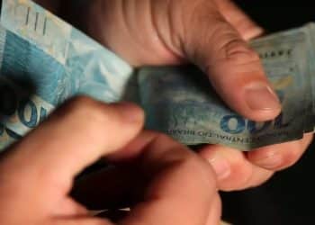 Mãos contam notas de R$ 100, referência ao piso de R$ 3.242 pago em 2026 ao técnico em radiologia, valor equivalente a dois salários mínimos.