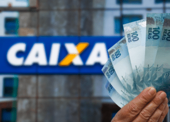 Mão segura notas de cem reais em frente ao letreiro da Caixa Econômica Federal, simbolizando as regras do ressarcimento, o dinheiro esquecido do fundo PIS/Pasep.
