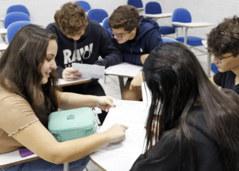 Imagem ilustrativa de cinco jovens sentados em carteiras escolares, debruçados sobre folhas de estudo, referência aos candidatos que se preparam para as atualidades do Enem