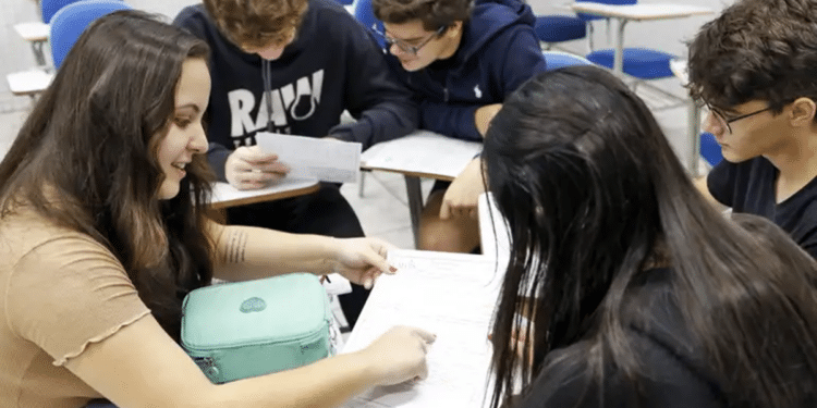 Imagem ilustrativa de cinco jovens sentados em carteiras escolares, debruçados sobre folhas de estudo, referência aos candidatos que se preparam para as atualidades do Enem