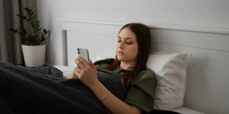 Jovem deitada na cama usando o celular, representando o uso excessivo das redes sociais e seus impactos no bem-estar.