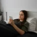 Jovem deitada na cama usando o celular, representando o uso excessivo das redes sociais e seus impactos no bem-estar.