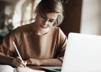 Jovem estudando concentrada ao lado de notebook em ambiente aconchegante.