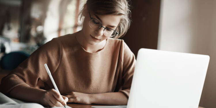 Jovem estudando concentrada ao lado de notebook em ambiente aconchegante.