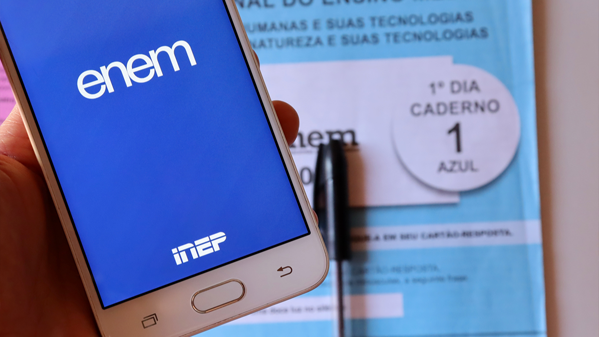 Celular aberto com tela azul e nome Enem destacado e prova no fundo.