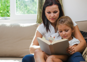 Mãe ensina filha a ler em casa durante momento de leitura no sofá.